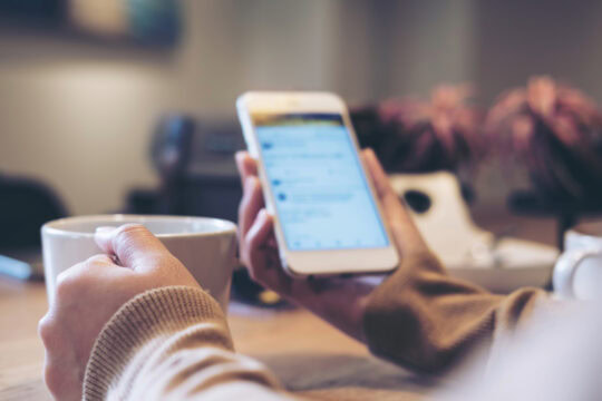 Closeup of a person holding both a mug of coffee and their cell phone. Their phone is open to a social media account.
