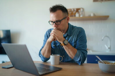 A middle-aged white man with glasses sits at his kitchen table looking at his laptop screen. He looks tense, with his elbows on the table and his hands clasped in front of his mouth,.