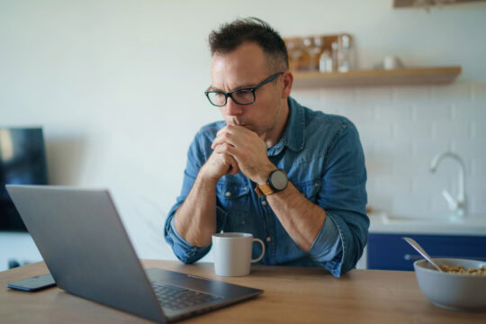 A middle-aged white man with glasses sits at his kitchen table looking at his laptop screen. He looks tense, with his elbows on the table and his hands clasped in front of his mouth,.