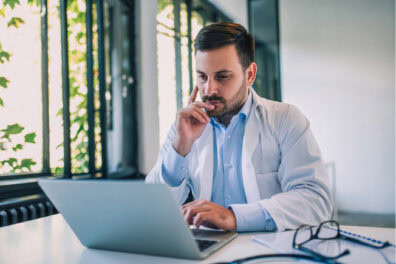 A doctor works on his laptop with a thoughtful look on his face.
