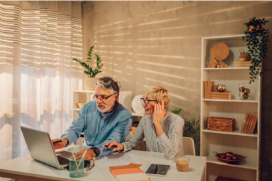 An older couple sit at a desk together and work on their finances. on the table in front of them are notebooks, a tablet, and a laptop.