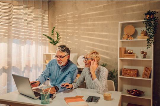 An older couple sit at a desk together and work on their finances. on the table in front of them are notebooks, a tablet, and a laptop.
