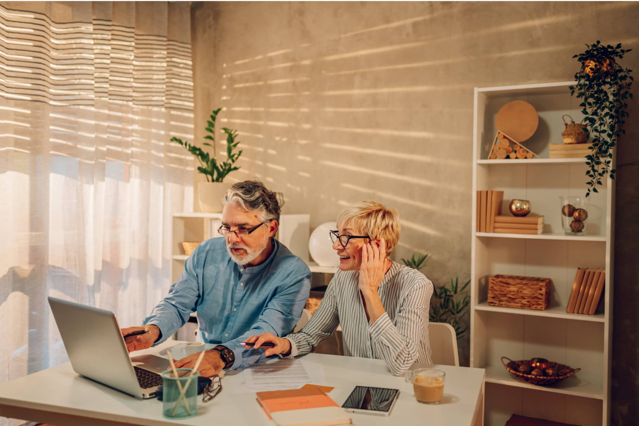An older couple sit at a desk together and work on their finances. on the table in front of them are notebooks, a tablet, and a laptop.