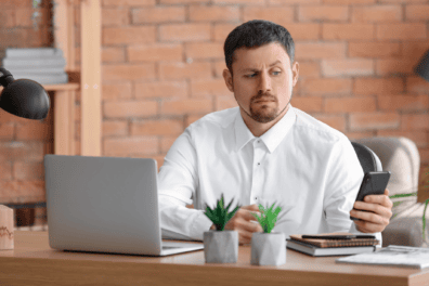 A man at work checks his phone with a suspicious look on his face. He's wearing a button up shirt and is sitting at his desk at work.
