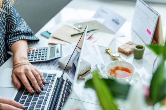 Close up of a woman's workspace. It's a cluttered desk with a laptop, tea, and lots of papers scattered about.
