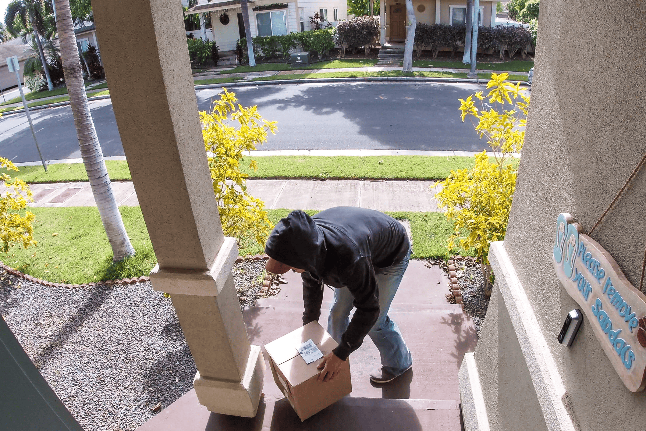 A view of a home's front porch from near the roof, as with a security camera. In frame is a hooded figure taking a package off of the doorstep.