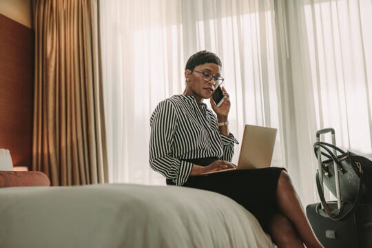 A Black businesswoman takes a call and works on her laptop from her hotel room. Her luggage is unopened next to her.