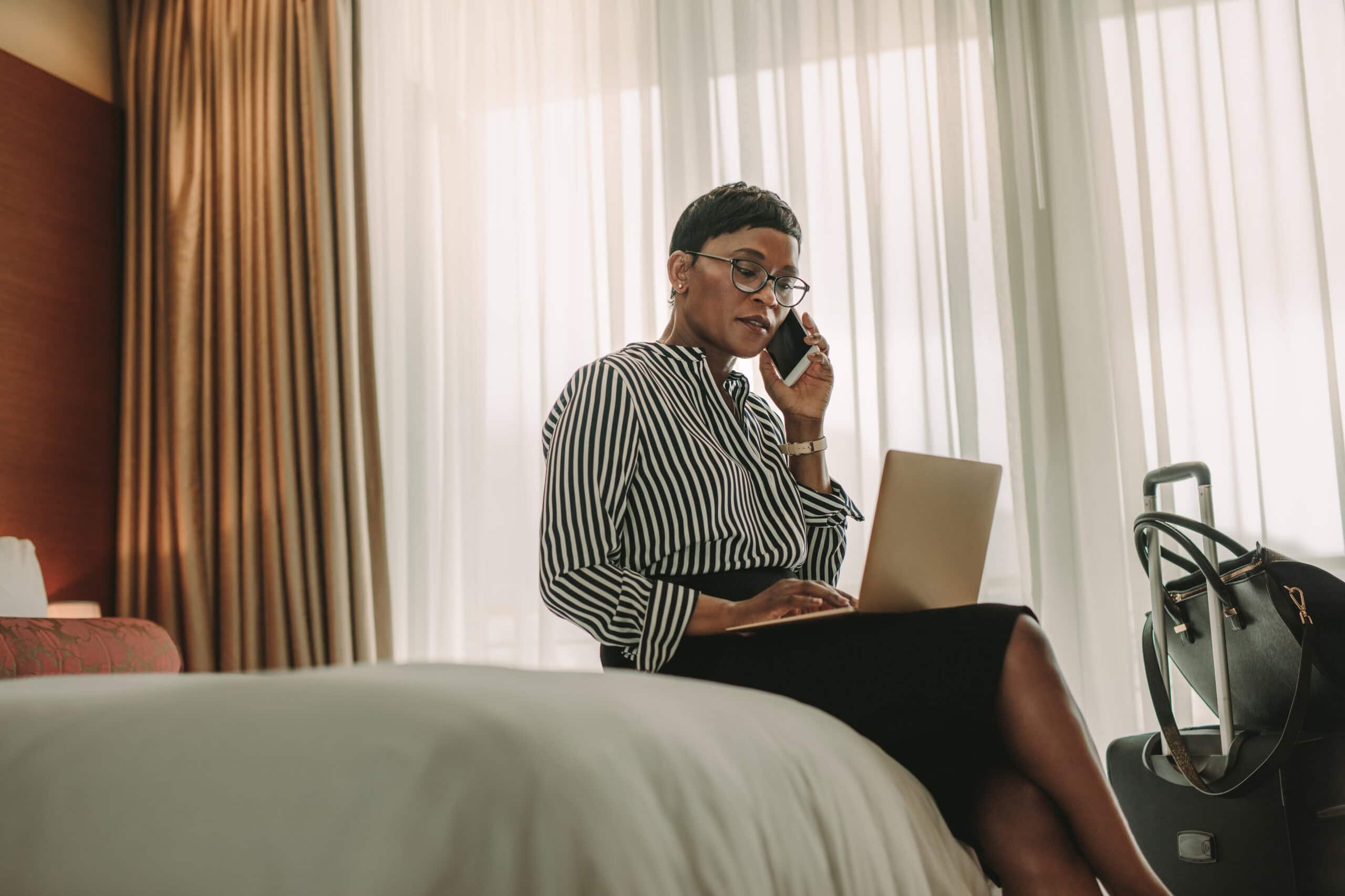 A Black businesswoman takes a call and works on her laptop from her hotel room. Her luggage is unopened next to her.