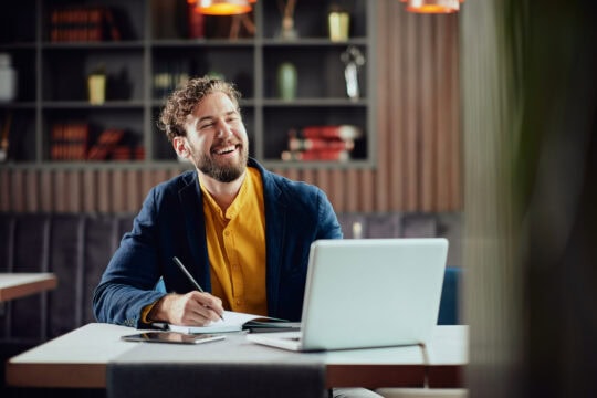 A white man with glasses and a beard smiles as he takes notes while working on his laptop.