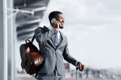 A Black man in a suit talks on the phone while waiting for a ride outside of the airport.