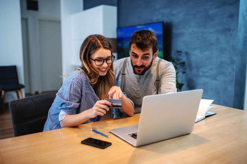 Smiling couple talking about things they want to buy online looking at a laptop on the dining table. The woman is holding and pointing at credit card.