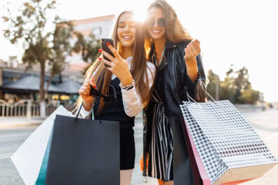 Young women in sunglasses hold shopping bags and use a cell phone while smiling and standing on a city street.