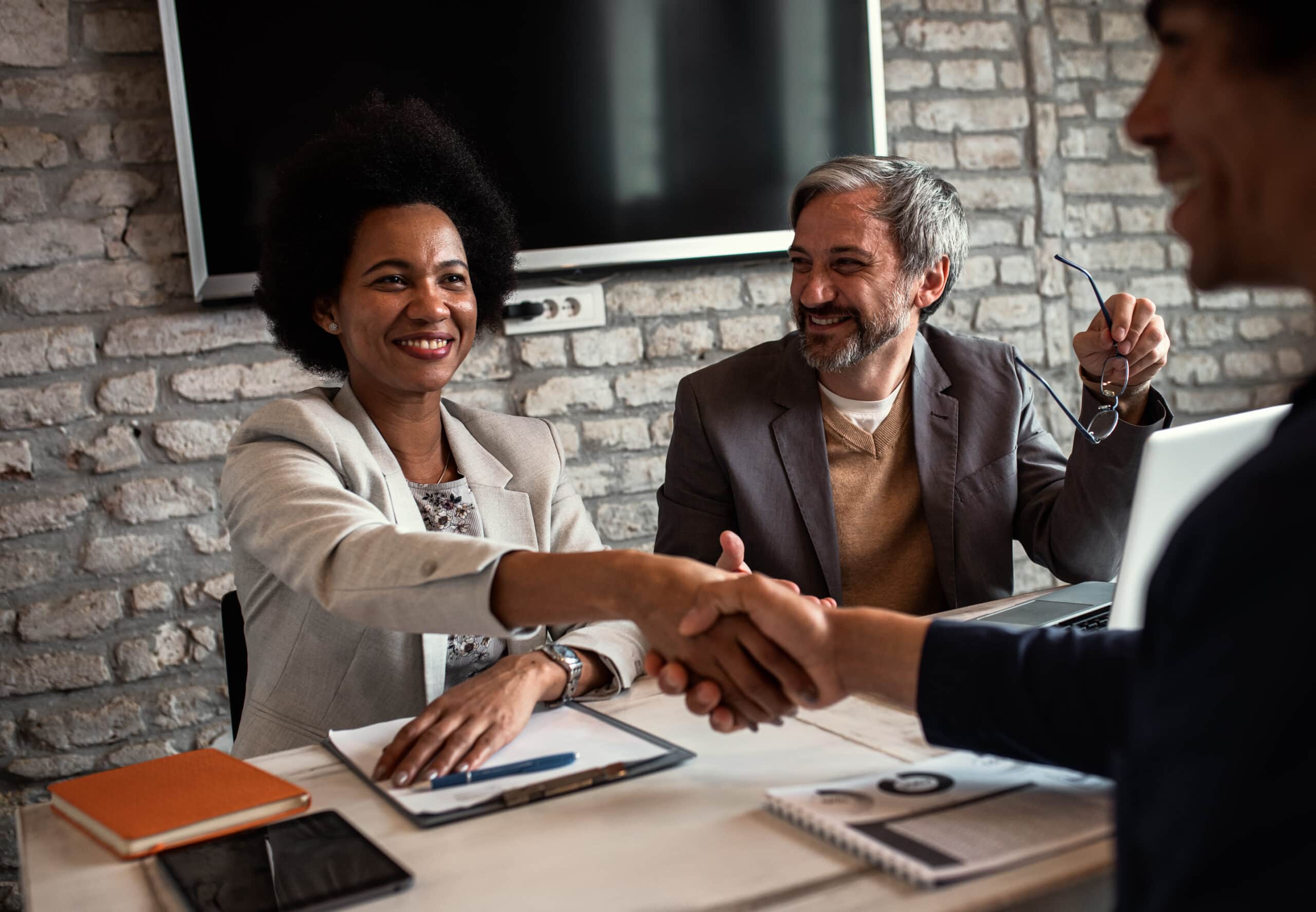 A middle aged Black woman shakes hands with a new hire she and the white middle aged man sitting next to her just interviewed.