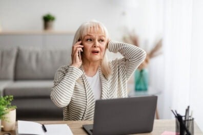A senior woman sits at a table with her laptop in front of her. She's on the phone and she has a shocked and worried look on her face.