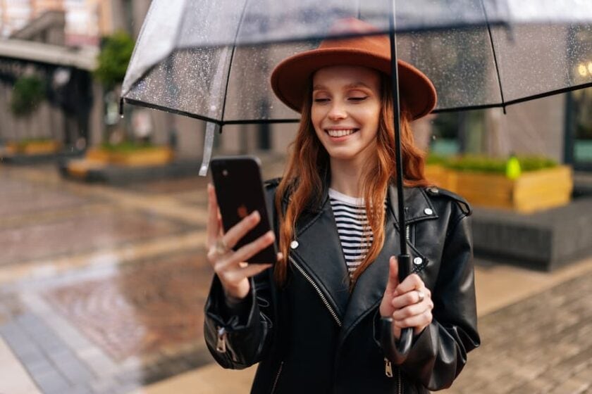 Portrait of pretty cheerful young woman tourist holding transparent umbrellain one hand and phone in the other standing in city in autumn rain