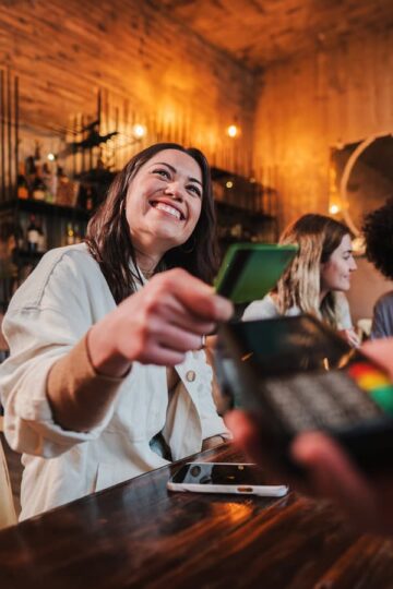 Vertical portrait of happy confident young woman paying the bill with her contactless Jeanne D'Arc MoneyStrong credit card in a restaurant.