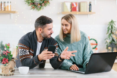 A husband and wife work on their budget together at home. They’re working on their laptop at their kitchen island surrounded by Christmas decorations.