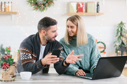 A husband and wife work on their budget together at home. They’re working on their laptop at their kitchen island surrounded by Christmas decorations.