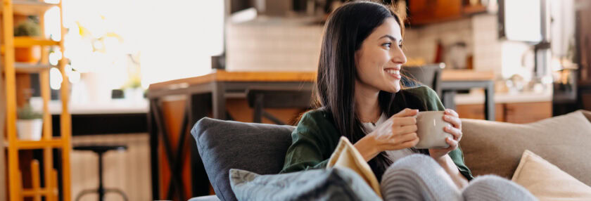 Young woman sitting on a comfortable sofa in a cozy living room, enjoying a warm cup of coffee or tea, smiling while gazing out the window, radiating happiness and serenity
