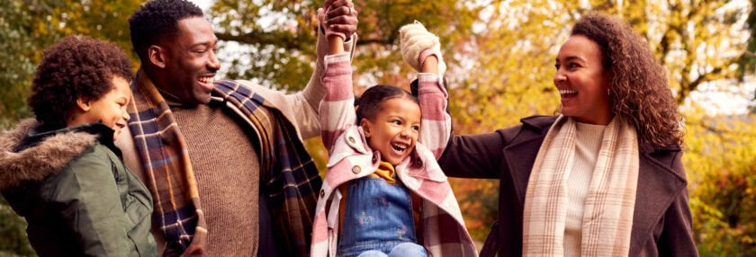 Smiling Family Having Fun Swinging Daughter On Walk Through Countryside Against Autumn Trees