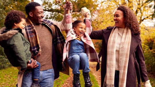 Smiling Family Having Fun Swinging Daughter On Walk Through Countryside Against Autumn Trees