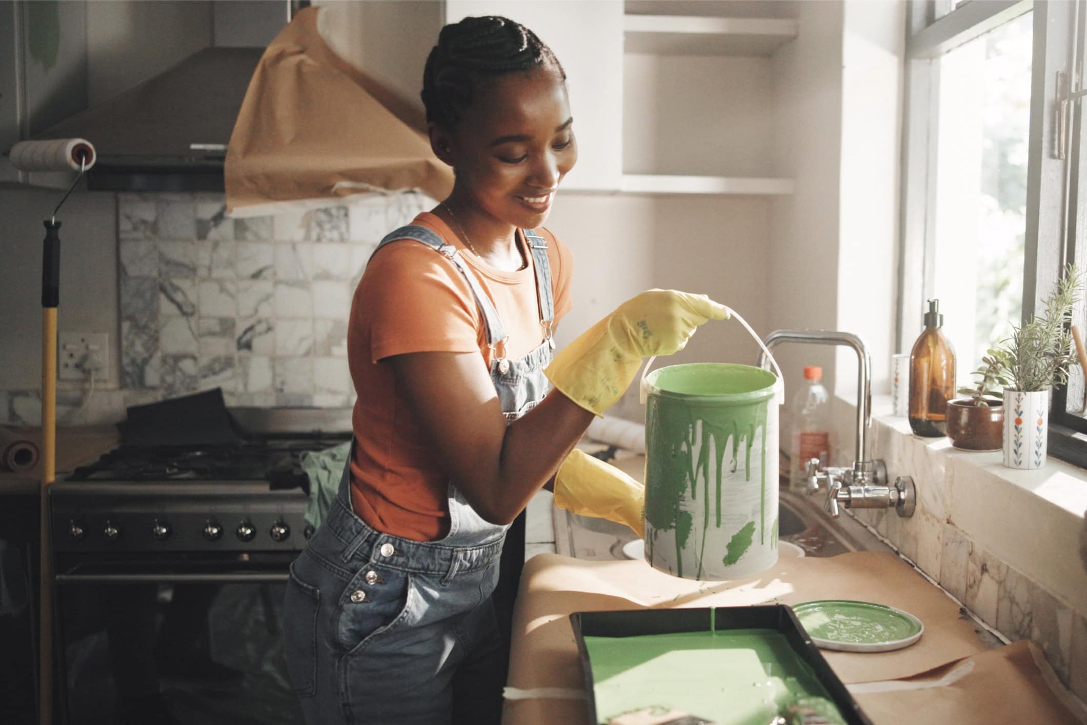 A Black woman wearing a t-shirt and overalls pours a gallon of paint into a tray as she gets ready to paint her kitchen.