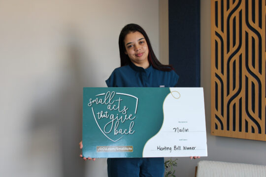 A woman holds a sign that reads "Small Acts That Give Back" and "My name is Nailin and I am a Heating Bill Winner"