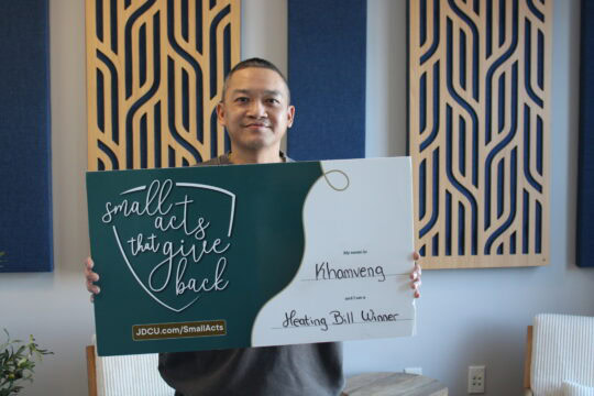 A woman holds a sign that reads "Small Acts That Give Back" and "My name is Khamveng and I am a Heating Bill Winner"