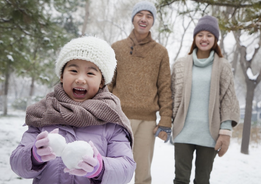 Young girl carrying snow balls in front of parents in park in winter