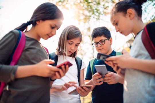A group of school children stand together looking at their smartphones.