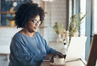 A Black woman with glasses and curly hair sits in a cafe working on her laptop.