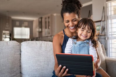 A mom and her daughter sit on the couch and play on a tablet together.