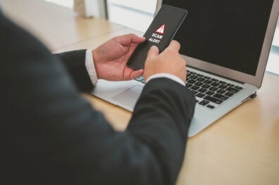 Closeup of businessman's hands as he sits in front of his laptop. He's holding his phone and there's a "Scam Alert" message on the screen.