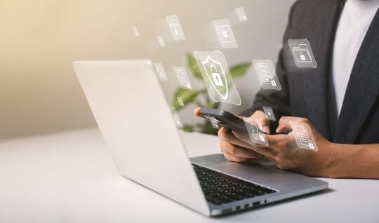 A person using their phone and laptop with icons of locked and secured files and information.