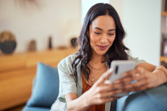 Happy, relaxed, beautiful woman using her smartphone while sitting on sofa at home.