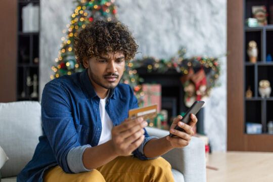 Hispanic man holding bank credit card and phone in his living room, which is decorated for Christmas.