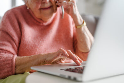 An older white woman talks to someone on the phone while using her laptop. The photo is cropped so you cannot see the top half of her face.