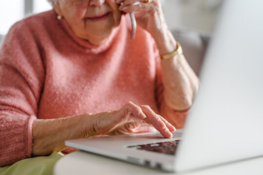 An older white woman talks to someone on the phone while using her laptop. The photo is cropped so you cannot see the top half of her face.