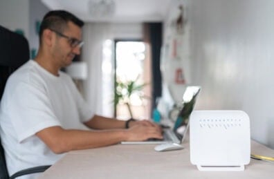 Closeup of a home internet router. In the background is a man with glasses working on a laptop.