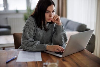 A brown-haired woman searches for a job online. She's sitting at a table at home with her laptop and resume in front of her.
