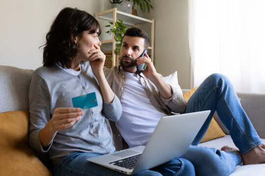 A couple sit on their couch and look worried. A man is on the phone looking at a woman, who has a credit card in one hand and the other over her mouth, and her laptop is on her lap.