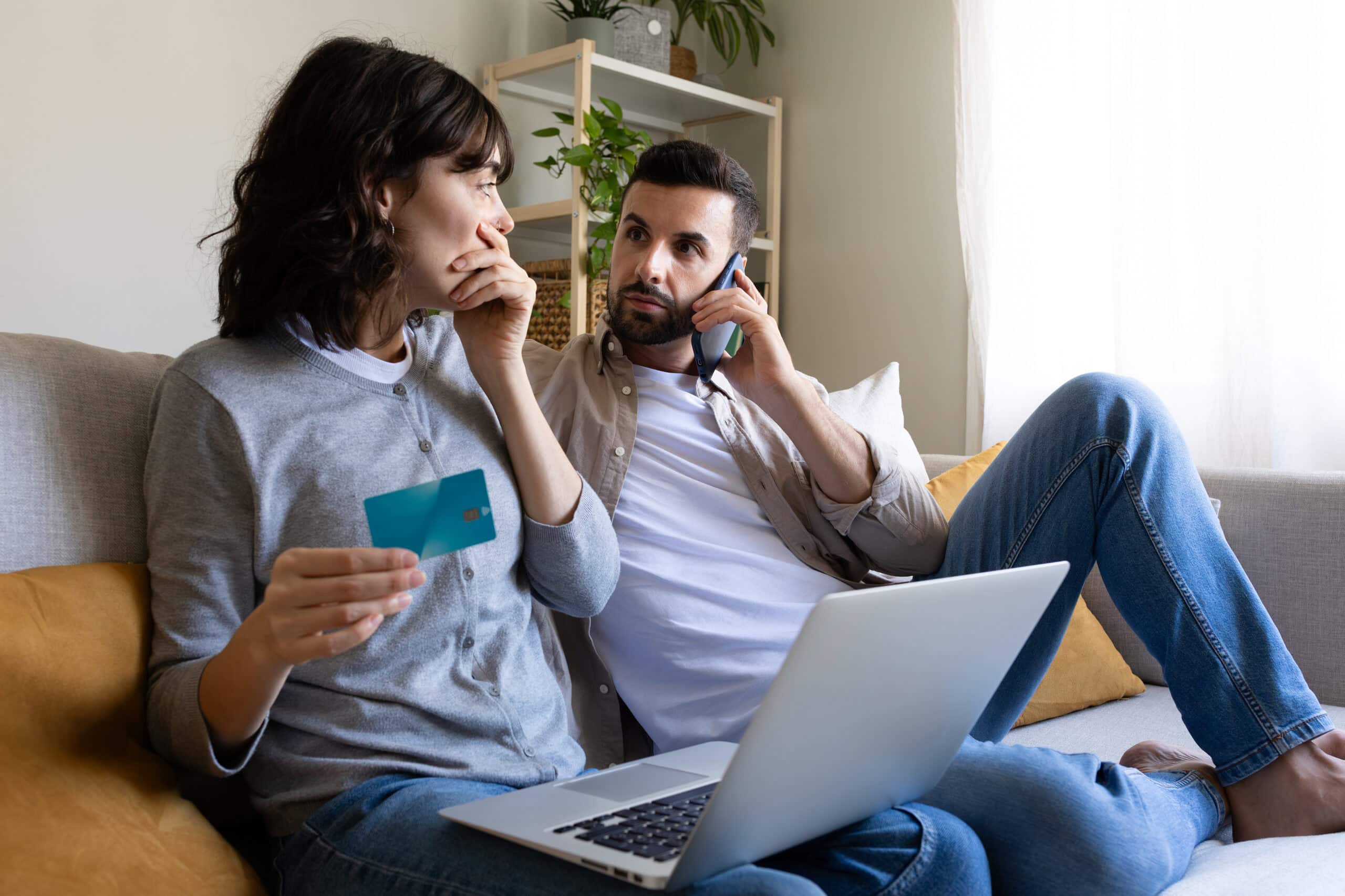 A couple sit on their couch and look worried. A man is on the phone looking at a woman, who has a credit card in one hand and the other over her mouth, and her laptop is on her lap.