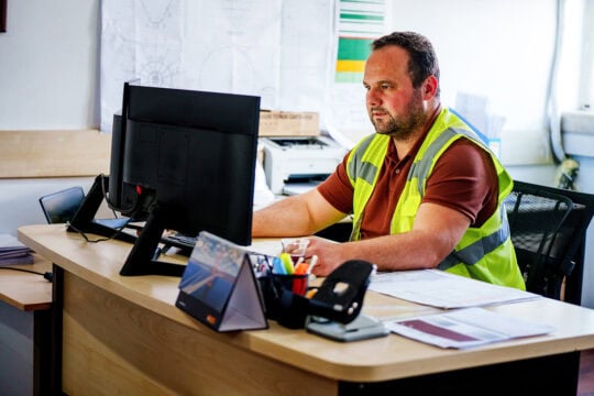 A man in a high-vis vest works on his computer at his office desk.
