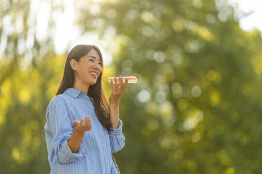 A cheerful young Asian woman smiles while using a voice assistant on her smartphone outside in a sunlit park.