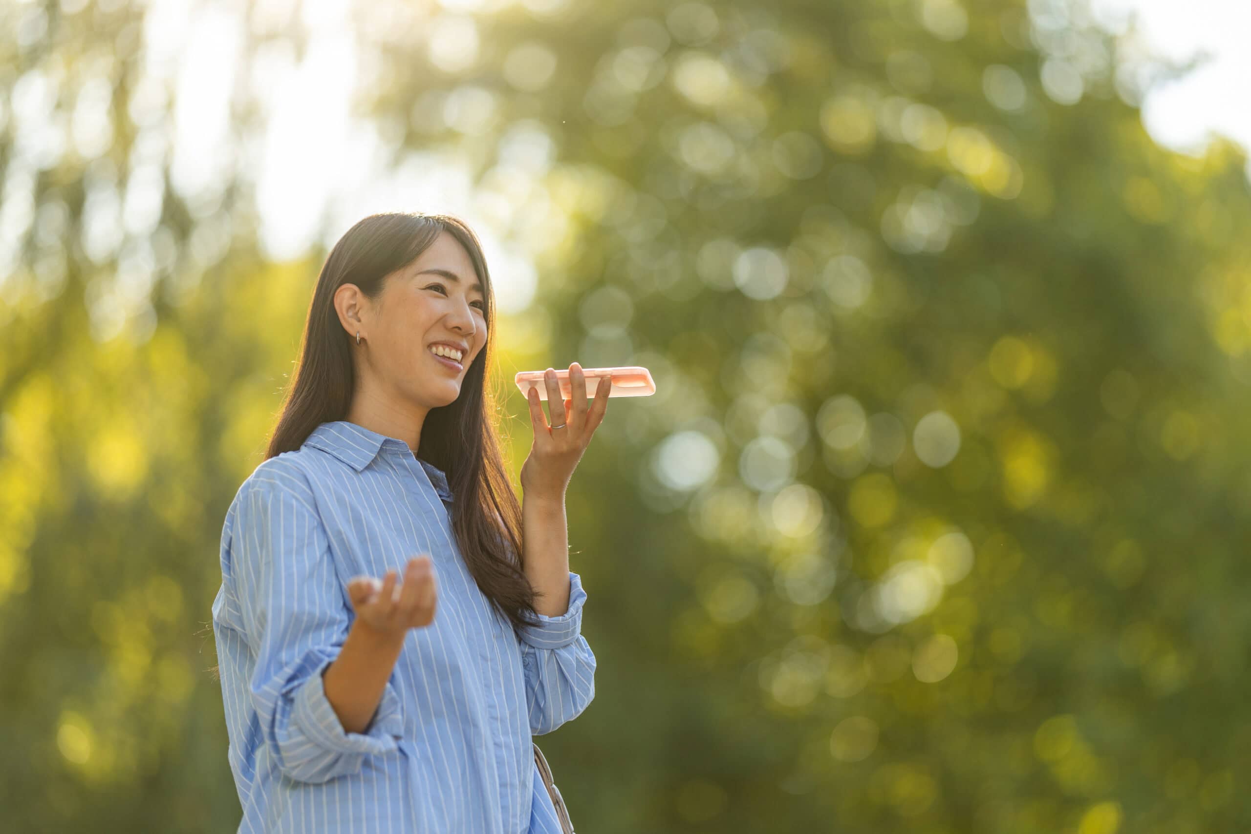 A cheerful young Asian woman smiles while using a voice assistant on her smartphone outside in a sunlit park.