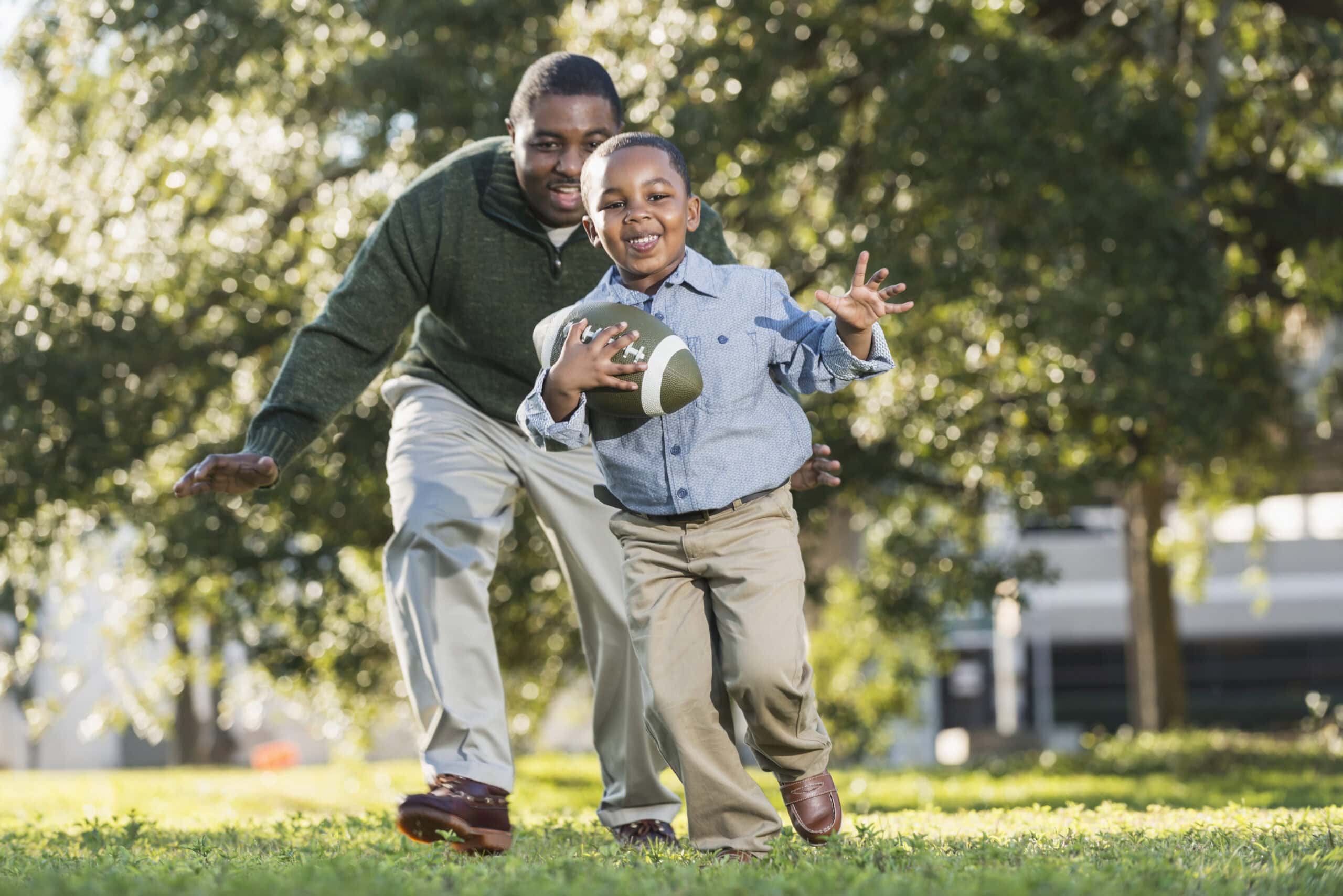 An African American man in his 40s playing football with his 5 year old son. It is a bright, sunny spring day. They are outdoors in the park or the yard, trees out of focus in the background. The boy is holding the ball, running, and his dad is chasing him from behind.