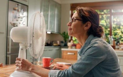 A woman sits at her kitchen table with an oscillating fan blowing in her face.